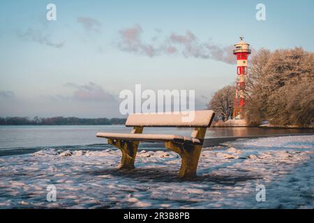 Germania, Amburgo, panchina vuota sulla riva innevata del fiume Elba con il faro Wittenbergen sullo sfondo Foto Stock