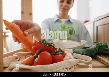 Donna che raccoglie carota fresca da pomodori in borsa riutilizzabile sul tavolo a casa Foto Stock