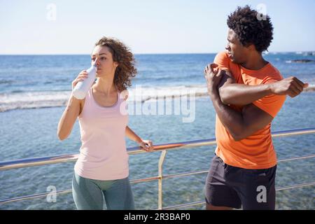 Uomo che si esercita con ragazza bere acqua da ringhiera in zona costiera Foto Stock