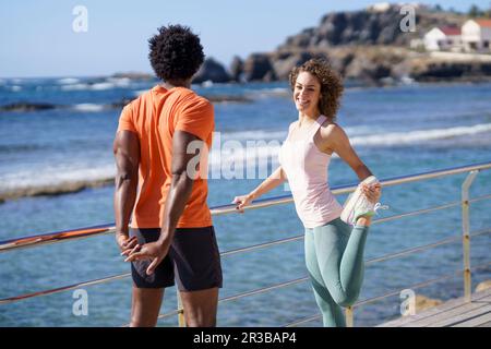 Donna sorridente che guarda il ragazzo fare esercizio stretching sul molo nella zona costiera Foto Stock