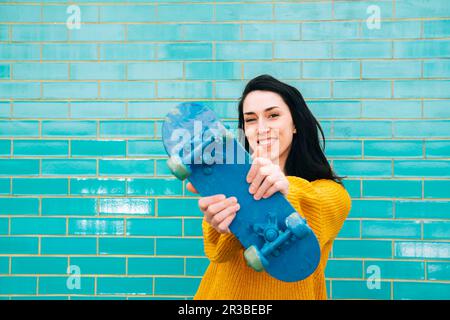 Donna sorridente in maglione che mostra skateboard blu di fronte al muro di mattoni Foto Stock