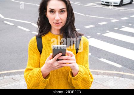 Giovane donna in maglione giallo con smartphone vicino alla strada Foto Stock