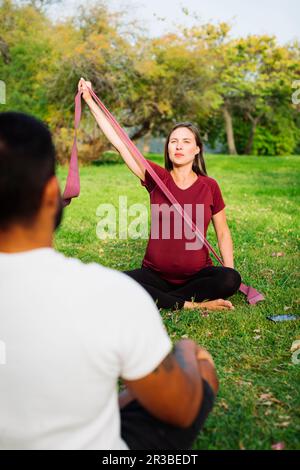 Donna incinta che tiene la fascia di resistenza seduta con l'uomo sul prato Foto Stock