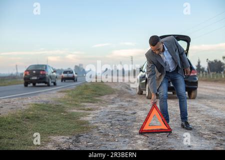 Giovane uomo latino che mette un triangolo di avvertimento dietro una macchina rotta con spazio di copia. Foto Stock