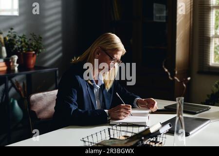 Psicologo biondo senior che prepara appunti alla scrivania in ufficio Foto Stock