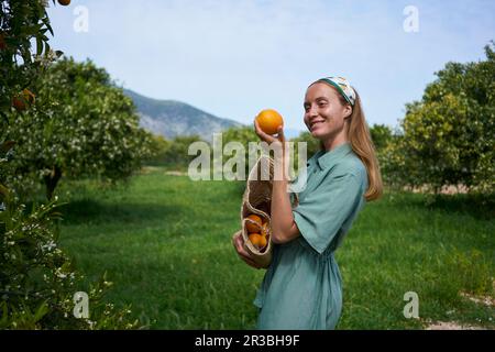 Giovane donna sorridente che tiene arancione in piedi nel frutteto Foto Stock