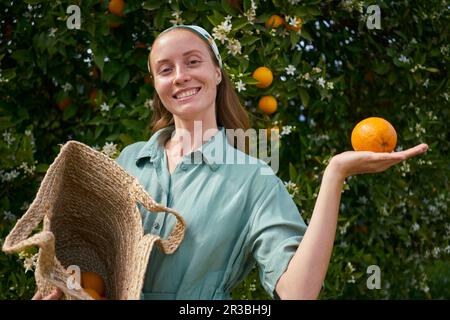 Giovane donna sorridente con arancio in mano vicino all'albero al frutteto Foto Stock