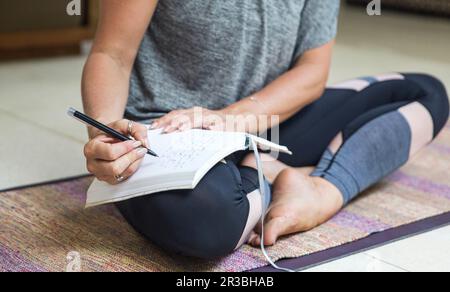 Donna che scrive in diario seduta a gambe incrociate sul tappeto di esercizio a casa Foto Stock