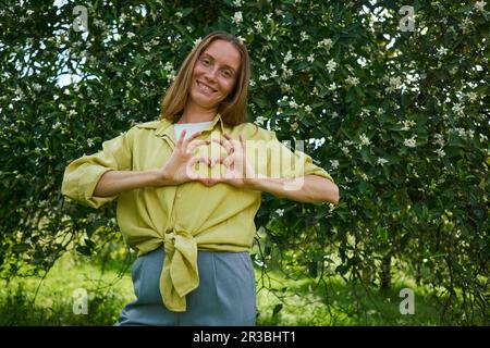 Donna che fa il gesto del cuore in piedi di fronte all'albero in giardino Foto Stock