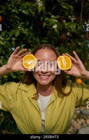 Donna sorridente che tiene fette di arance fresche in frutteto Foto Stock