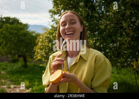 Donna felice con paglia di metallo riutilizzabile e fette di arancia al frutteto Foto Stock