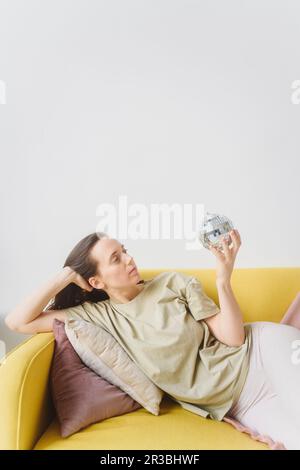 Donna con palla di vetro sdraiata sul divano giallo Foto Stock