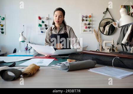 Giovane donna in abbigliamento da lavoro che guarda carte con disegni in officina, scegliendo nuovi modelli di abbigliamento da tavolo con pelle arrotolata Foto Stock