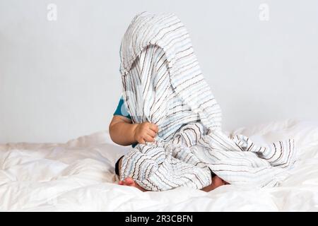 Piccolo ragazzo caucasico che gioca sotto una coperta a strisce su biancheria da letto bianca Foto Stock