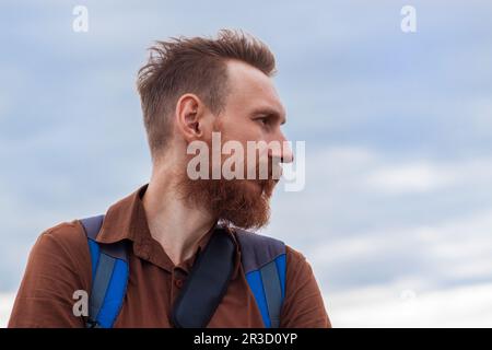 Bell'uomo di hippster con barba lussureggiante e baffi su sfondo cielo nuvoloso blu. Ragazzo rosso con zaino all'aperto. Vista laterale maschile seria e brutale. Foto Stock