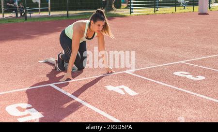 Giovane atleta femminile oltre la linea di partenza, assumendo una posizione di partenza accovacciata e correndo lungo una pista atletica dello stadio Foto Stock