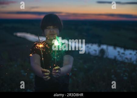 Un ragazzo carino tiene in mano gli scintillatori, fuochi d'artificio la sera d'estate al tramonto. Atmosfera festiva. Foto Stock