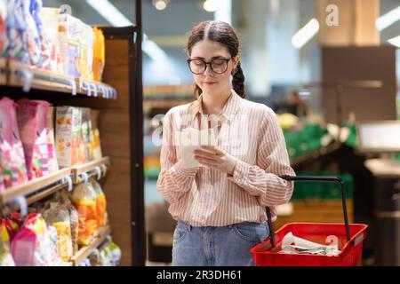 Ritratto di giovane donna caucasica bella indossare occhiali contiene la lista dei prodotti e scrivere in esso. Concetto di shopping in supermercato e consumismo. Foto Stock