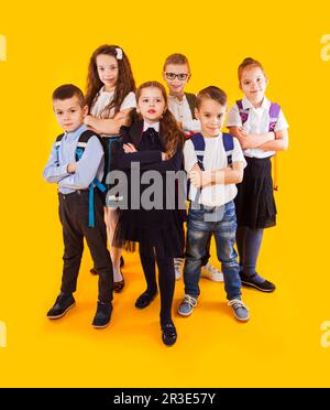 Gruppo di bambini felici in uniforme con borse da scuola abbracciando e guardando alla fotocamera su sfondo giallo Foto Stock