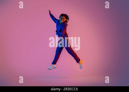 Ritratto a tutta lunghezza di donna nera eccitata in sportswear salta su sfondo rosa neon, spazio libero Foto Stock