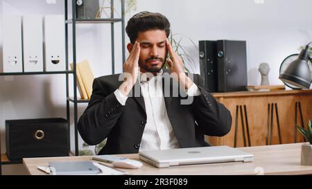 Uomo d'affari indiano malato stanco che soffre di mal di testa tensione problema ed emicrania, stress a casa scrivania ufficio posto di lavoro. L'uomo di lavoro di broker freelancer con bearded lavora su notebook. Lavoro eccessivo Foto Stock