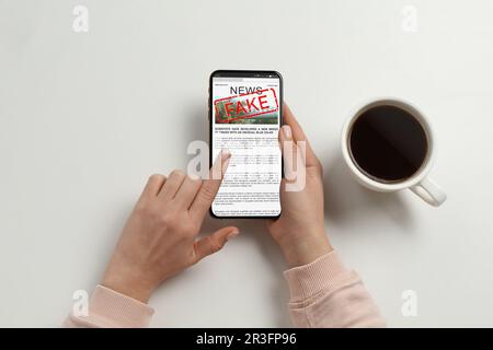 Informazioni false. Donna che utilizza il telefono cellulare per leggere le notizie al tavolo bianco, primo piano Foto Stock