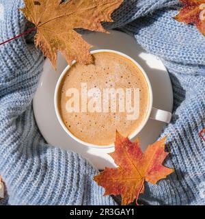 Tazza bianca di caffè mattutino caldo su maglione blu lavorato a maglia con sfondo giallo acero foglie. Concetto di casa accogliente. Estetica b Foto Stock