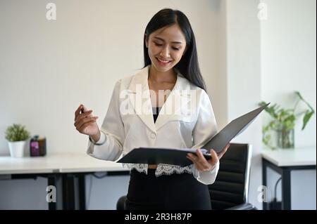 Giovane assistente o donna d'affari asiatica sorridente e affascinante che rivede documenti aziendali in una cartella nel suo ufficio. Foto Stock