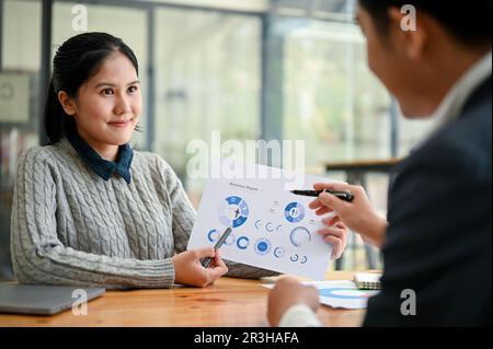 Una giovane lavoratrice asiatica sta parlando con il suo manager maschile dei rapporti aziendali in ufficio. Foto Stock