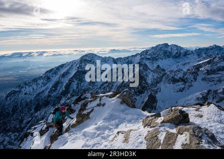 Avventuroso uomo escursionista in cima a una ripida scogliera rocciosa che domina inverno alpino come paesaggio moutain di alti Tatra, Slovacchia. Paesaggio alpino di montagna Foto Stock