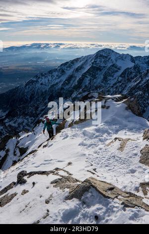 Avventuroso uomo escursionista in cima a una ripida scogliera rocciosa che domina inverno alpino come paesaggio moutain di alti Tatra, Slovacchia. Paesaggio alpino di montagna Foto Stock