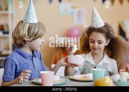 Ritratto di due bambini felici che indossano cappelli da festa e che si gustano dolci a tavola Foto Stock