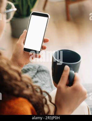 Vista a spalla di una donna che tiene in mano il caffè e lo smartphone con schermo cellulare bianco mock-up utilizzando le app mobili su un telefono cellulare. Cellulare Foto Stock