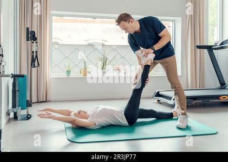 Personal trainer che assiste una donna di sessant'anni con un esercizio di stretching in palestra, è sdraiata su un tappetino con una gamba estesa in avanti e braccia bac Foto Stock