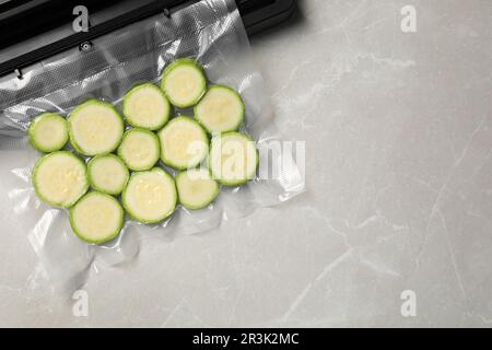 Sigillante per confezionamento sottovuoto e sacchetto di plastica con zucchine tagliate su tavolo in marmo grigio chiaro, vista dall'alto. Spazio per il testo Foto Stock