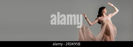 elegante donna incinta con capelli ondulati in posa in chiffon drappeggi e accessori dorati isolati su sfondo grigio, elegante gravidanza Foto Stock