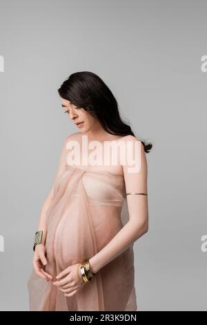 donna incinta aggraziata con capelli brunette ondulati che posano con panno in chiffon beige e braccialetti dorati isolati su sfondo grigio, co moda maternità Foto Stock