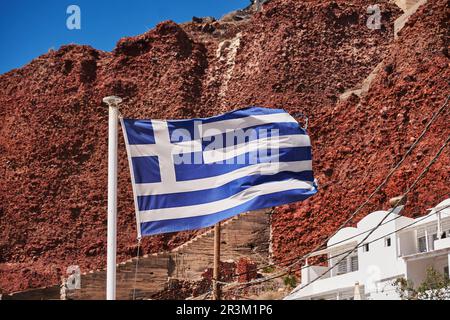 Bandiera greca a Amoudy Bay Red Volcanic Cliffs - Oia Village - Santorini Island, Grecia Foto Stock