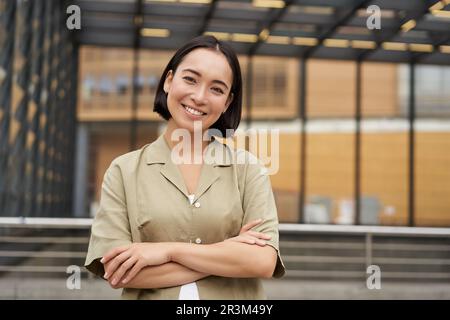 Ritratto di giovane donna asiatica in piedi con fiducia, braccia incrociate sul petto e sorridente, in posa all'aperto sulla strada Foto Stock