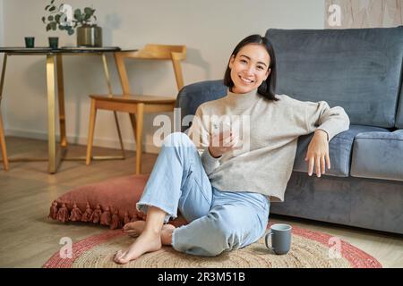 Ritratto di donna coreana elegante siede sul pavimento con lo smartphone, utilizzando il telefono cellulare, sorridendo soddisfatto, concetto di rimanere a casa Foto Stock