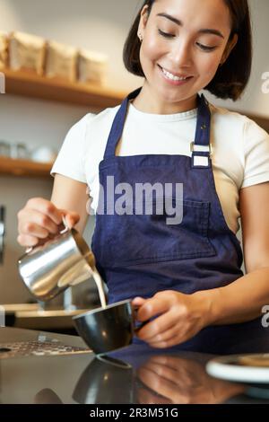 Shot verticale di barista ragazza asiatica brunette, con grembiule, caffè, preparazione dell'ordine in caffetteria, versamento del latte in tazza per il cappu Foto Stock