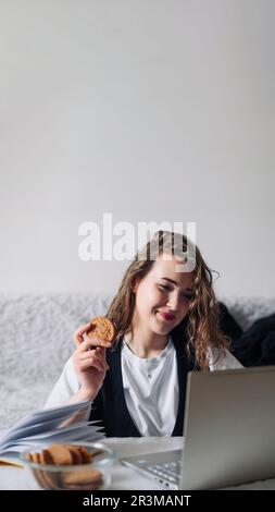 Giovane donna redhead guardando serie o podcast guardando lo schermo con interesse tenere cookie in mano seduto al tavolo da cucina con vaso di vetro e. Foto Stock