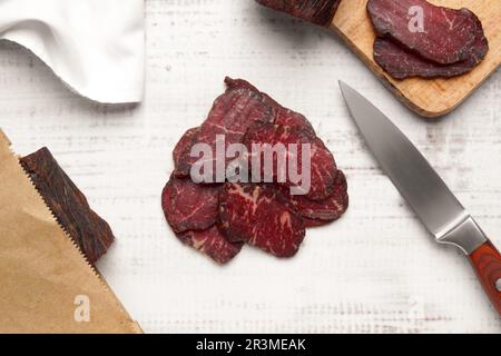 Pezzi di delizioso manzo a scatti e coltello su tavolo di legno bianco, piatto Foto Stock