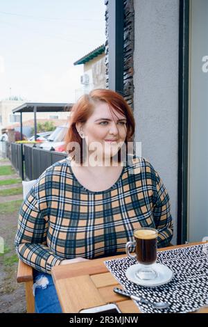 giovane donna latina di più dimensioni con capelli rossi è seduta fuori dalla caffetteria sorridendo guardando dritto in avanti, prestando attenzione alla chat del suo amico. vert Foto Stock