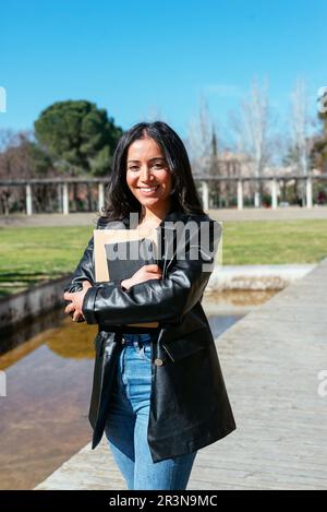Giovane studentessa ispanica positiva in giacca di pelle sorridente e guardando la macchina fotografica mentre si è in piedi con libri a braccia incrociate in luce del giorno in città p Foto Stock