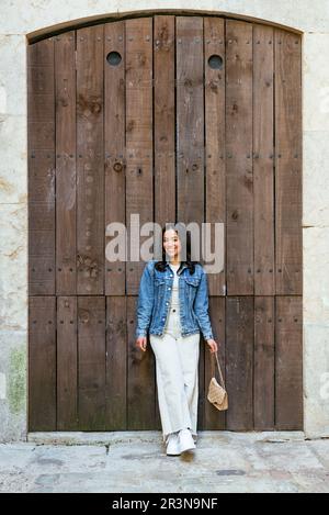 Corpo pieno di positiva giovane donna ispanica con borsa sorridente e guardando la macchina fotografica mentre si appoggia sulla porta di legno del vecchio edificio in luce del giorno Foto Stock