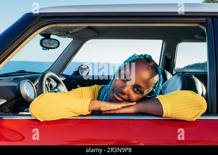 Vista frontale di una giovane ragazza afroamericana sorridente in capelli nuziali blu seduta sul sedile anteriore dell'auto mentre si guarda la macchina fotografica e si appoggia con le mani sul wi Foto Stock