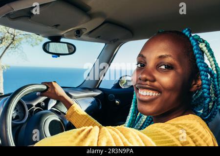 Ritratto di allegra driver afro-americana in capelli intrecciati blu seduti in auto con la mano sul volante e guardando la fotocamera contro nuvoloso Foto Stock