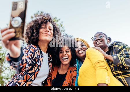 Da sotto gruppo di felici amici multietnici in abiti eleganti sorridendo mentre si prende selfie in piedi sotto il cielo blu luminoso nel parco di giorno Foto Stock