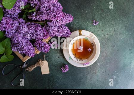 Gustoso tè nero in tazza bianca vintage su tavolo in cemento verde menta con fiori di lilla aromatica Foto Stock